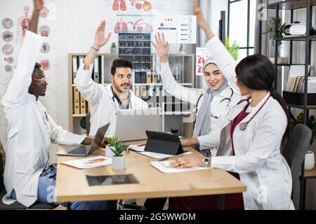 Team of four international doctors holding hands up for expressing successful cooperation about various medical moments. Health care workers sitting together at tablet and using modern gadgets. Stock Photo