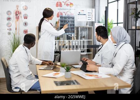 Group of four multiracial doctors using modern gadgets while examining tomography of patient during consilium. International colleagues sitting at room discussing various way of treatment. Stock Photo