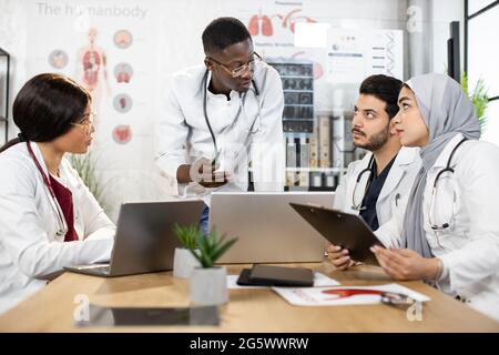 Four diverse medical workers in uniform having discussion at office with modern gadgets on table. Health care and teamwork concept. Stock Photo