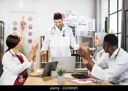 Multiracial students in white lab coats sitting at interactive classroom and clapping in the end of interesting lecture. Arabian male doctor using modern laptop for educational process. Stock Photo