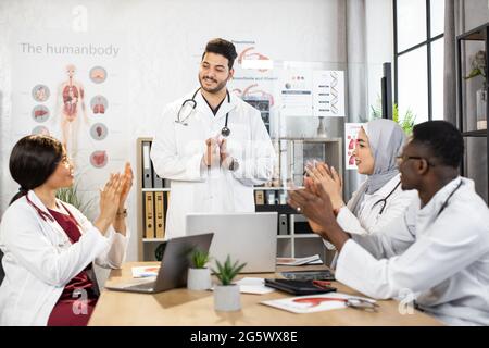 Arabic male scientist standing at interactive classroom and leading lecture for diverse colleagues. Multiethnic young people giving applause in the end of lesson. Concept of education and medicine. Stock Photo
