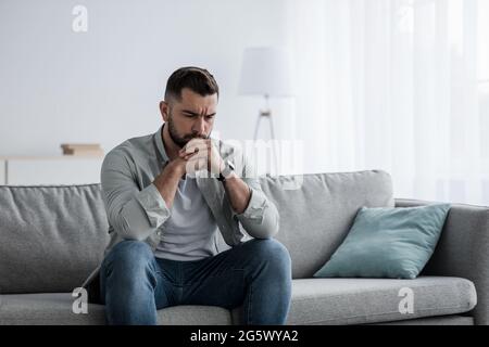 Thoughtful young serious handsome man sit on sofa at home, lost in thoughts, thinking about problem solving Stock Photo