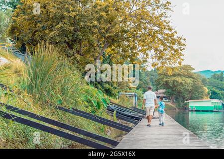 Back view of Asian grandfather and grandchild wear protective face mask to prevent Coronavirus (COVID-19) walking in a nature path on wooden bridge al Stock Photo