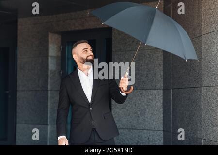 Mature office worker wearing classical suit holding umbrella while walking outdoors, going home after work in office Stock Photo