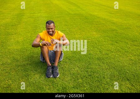Joyful young afro american sportsman in trendy sportswear sitting on the stadium grass, having rest break after football match, work out alone Stock Photo