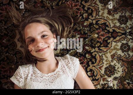 Smiling girl laying on the floor on a Persian carpet in a white dress. Stock Photo