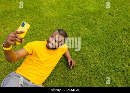 Joyful handsome afro american male laying on the grass, relaxing and taking a break after sport work out. Smiling black guy talking online with Stock Photo