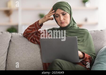 Concerned Middle-Aged Muslim Lady Using Laptop Working Online At Home Stock Photo