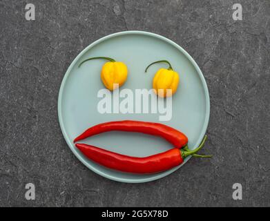 Plan view of red and yellow chilli peppers arrange like a face on a duck egg blue plate. Stock Photo