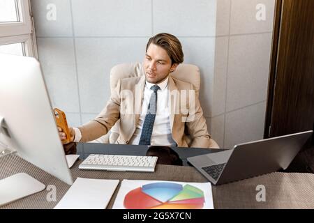 Young handsome businessman or ceo in trendy suit sitting at desk with laptop and PC, analyzing financial and marketing project report on phone and Stock Photo