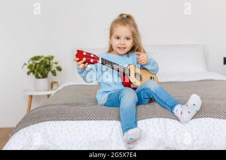 Cute little girl playing ukulele and singing it at home. 3 years old kid learning guitar. Concept of early childhood education, music hobby, talent, h Stock Photo