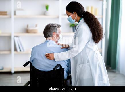 Young female doctor in face mask listening to old man's breathing, using stethoscope at home Stock Photo