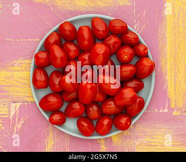 Plan view of small plum tomatoes on a duck egg blue plate, on a pink and yellow, rustic background Stock Photo