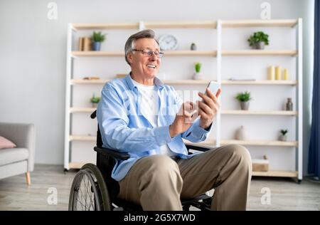 Happy disabled older man in wheelchair using cellphone, browsing web or watching movie at retirement home Stock Photo
