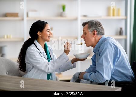 Caregiver feeding elderly man in wheelchair at home. Medical assistance for senior people with partial paralysis Stock Photo