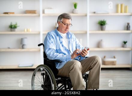 Senior disabled man in wheelchair using smartphone, checking messages, watching video, browsing web at home Stock Photo