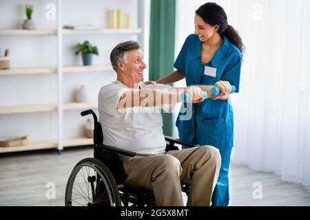 Female physiotherapist helping elderly man in wheelchair do exercises with dumbbells at health centre Stock Photo