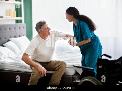 Caregiver helping elderly male to get into wheelchair at home. Rehabilitation and medical care for disabled seniors Stock Photo