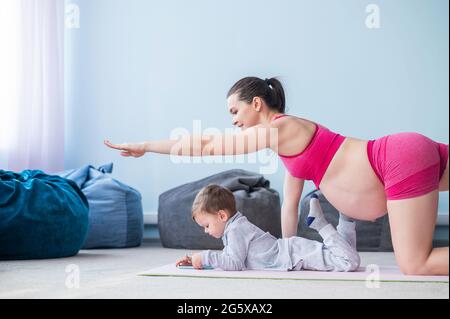 A pregnant woman in sportswear is doing gymnastics on a mat with her son. Girl on yoga with her child. Fitness mom and baby. Stock Photo