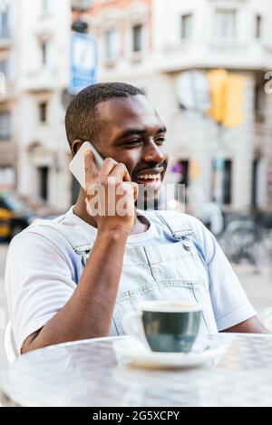 vertical portrait of an afro american young man. He is talking by phone and smiling at a bar terrace Stock Photo
