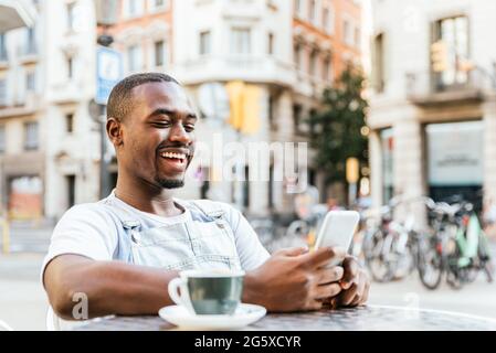 Attractive afro american man sitting at a bar terrace. He is using his phone and smiles. Stock Photo