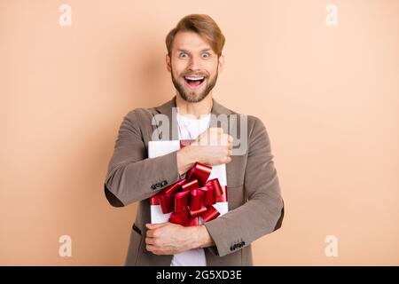 Photo of impressed funny young guy wear jacket smiling hugging gift box open mouth isolated beige color background Stock Photo