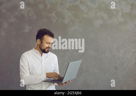 Young smiling man with a beard standing holding and typing on a laptop. Stock Photo