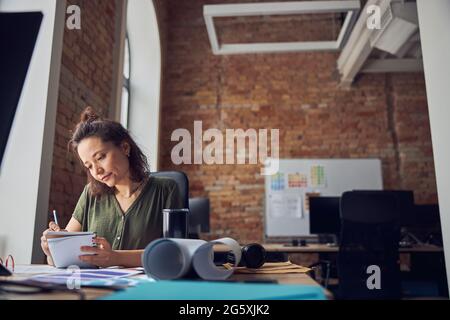Busy woman interior designer making notes in her notebook while working on new creative project, sitting at the table in her office Stock Photo