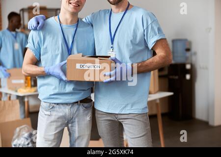 Cropped shot of two caucasian guys, male volunteers in blue uniform, protective gloves posing together and holding cardboard box for Donation Stock Photo