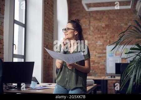 Smiling creative woman, interior designer or architect wearing glasses looking away, working on a blueprint for new project, standing in her office Stock Photo