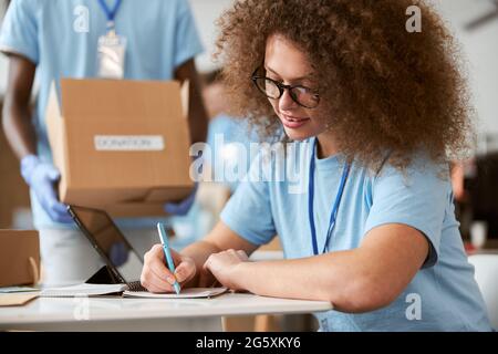 Young woman volunteer making notes and guy carrying cardboard box in the background while working on donation project indoors Stock Photo