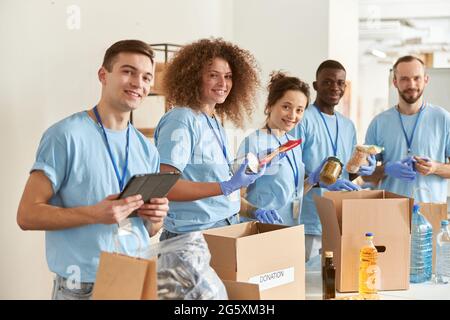 Portrait of happy volunteers smiling at camera while sorting, packing foodstuff in cardboard boxes, working together on donation project indoors Stock Photo