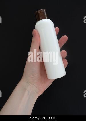 Female hand holding white cosmetic bottle on black background. Mock up, copy space. Stock Photo