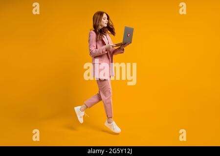 Red haired woman run jump typing laptop wearing elegant pink suit isolated on yellow background in studio, hurry up. side view portrait of lady workin Stock Photo