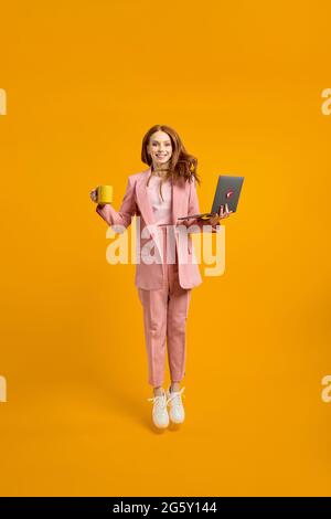 Full length profile photo of funny business lady jump high up hold laptop and cup of coffee in hands addicted worker always online wear pink suit outf Stock Photo