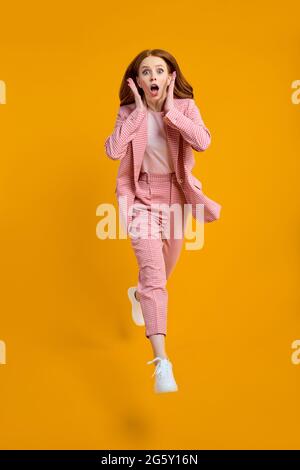 Full size photo of young happy shocked amazed surprised woman jumping in excitement isolated on yellow color background, wearing pink suit, raising ha Stock Photo