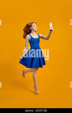 Full length body size photo of stylish trendy redhead woman taking selfie jumping in blue dress isolated yellow studio background, portrait. copy spac