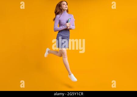 Dreamy redhead lady at weekend jumping, hold notebooks in hands on way home, in air, wearing casual shirt and skirt isolated yellow orange background. Stock Photo