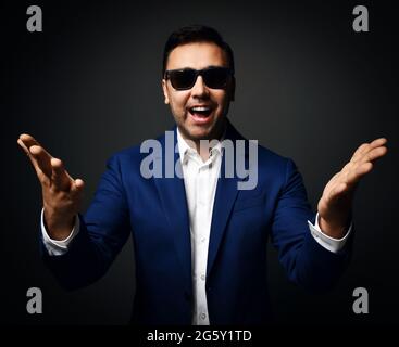 Happy laughing brunet man in blue jacket, white shirt and sunglasses stands holding hands with open palms up at camera