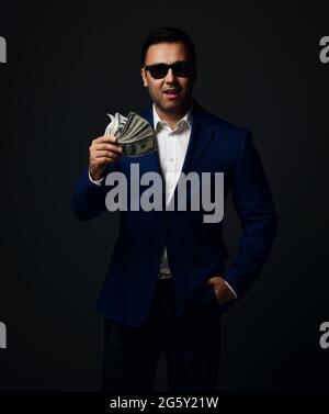 Portrait of insolent brunet businessman in blue jacket and sunglasses standing with fan of dollars cash in hand
