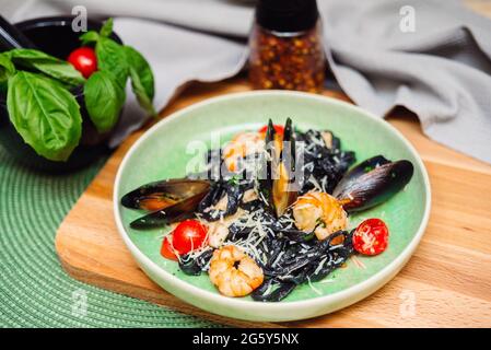 Black fettuccine pasta with shrimp, oysters, parmesan cheese, cherry tomatoes in a white wine sauce on a green plate and a wooden board on a backgroun Stock Photo