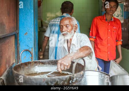 Elderly white bearded Indian male stirs contents in metal food pot as young lad watches from behind, Trichy, Tamil Nadu, India Stock Photo