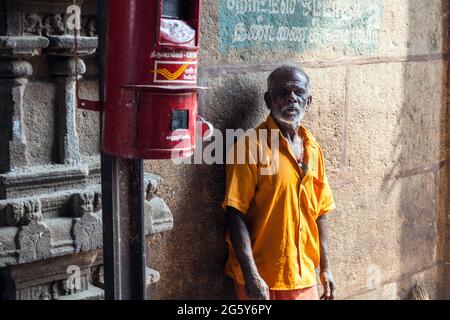 Portrait of sorrowful Indian male wearing orange with grey beard stood leaning against wall beside red India Post mailbox, Trichy, Tamil Nadu, india Stock Photo