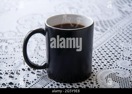 Steaming hot cup of black coffee in a black coffee mug on white lace tablecloth.  Classic drink still life with copy space on the black coffee cup. Stock Photo