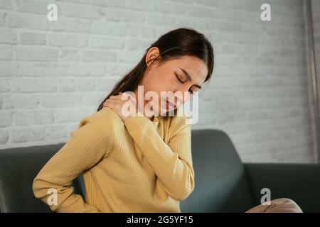 Asian young woman suffering from shoulder pain while sitting and resting on couch at home in casual clothing with closed eyes Stock Photo