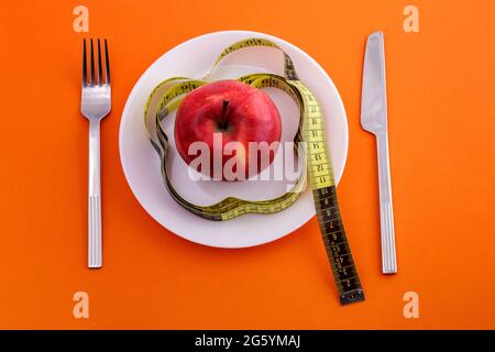 red apple lying on a white plate with a rewound measuring tape, knife and fork on an orange background, top view. Diet and healthy food concept Stock Photo