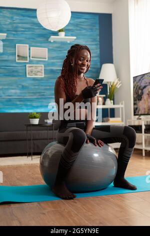 Sporty athletic black woman relaxing on stability ball checking social media using smartphone after intense workout exercising, in home living room sitting on fitness mat for muscle strenght. Stock Photo