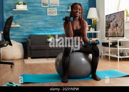 Joyful athletic black person in leggings using stability ball for training biceps, doing curls with dumbbells. Strong athletic person doing sports at home using modern equipment for Stock Photo