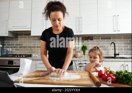 Woman kneading dough at home in the kitchen, next to daughter sitting on a chair and watching the cooking process. Family spending time concept. Teach Stock Photo