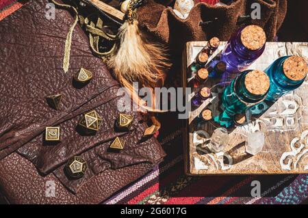 Overhead image of glass stopper bottles with colored liquid on a wooden box, metallic RPG gaming dice on a leather-bound book and part of a leather di Stock Photo